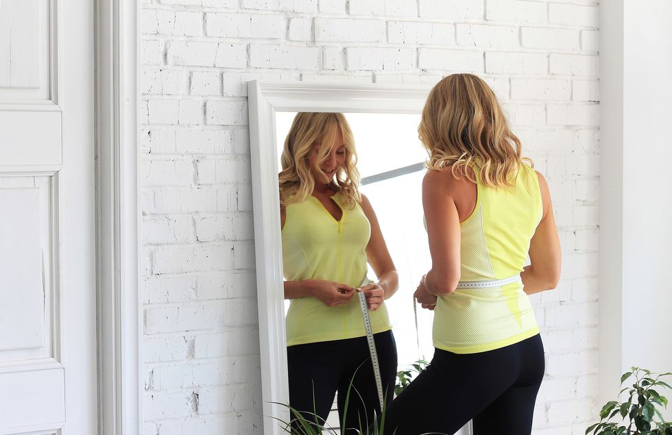 stay in shapeyoung woman with athletic body measures her waist with a measure type in front of a mirror