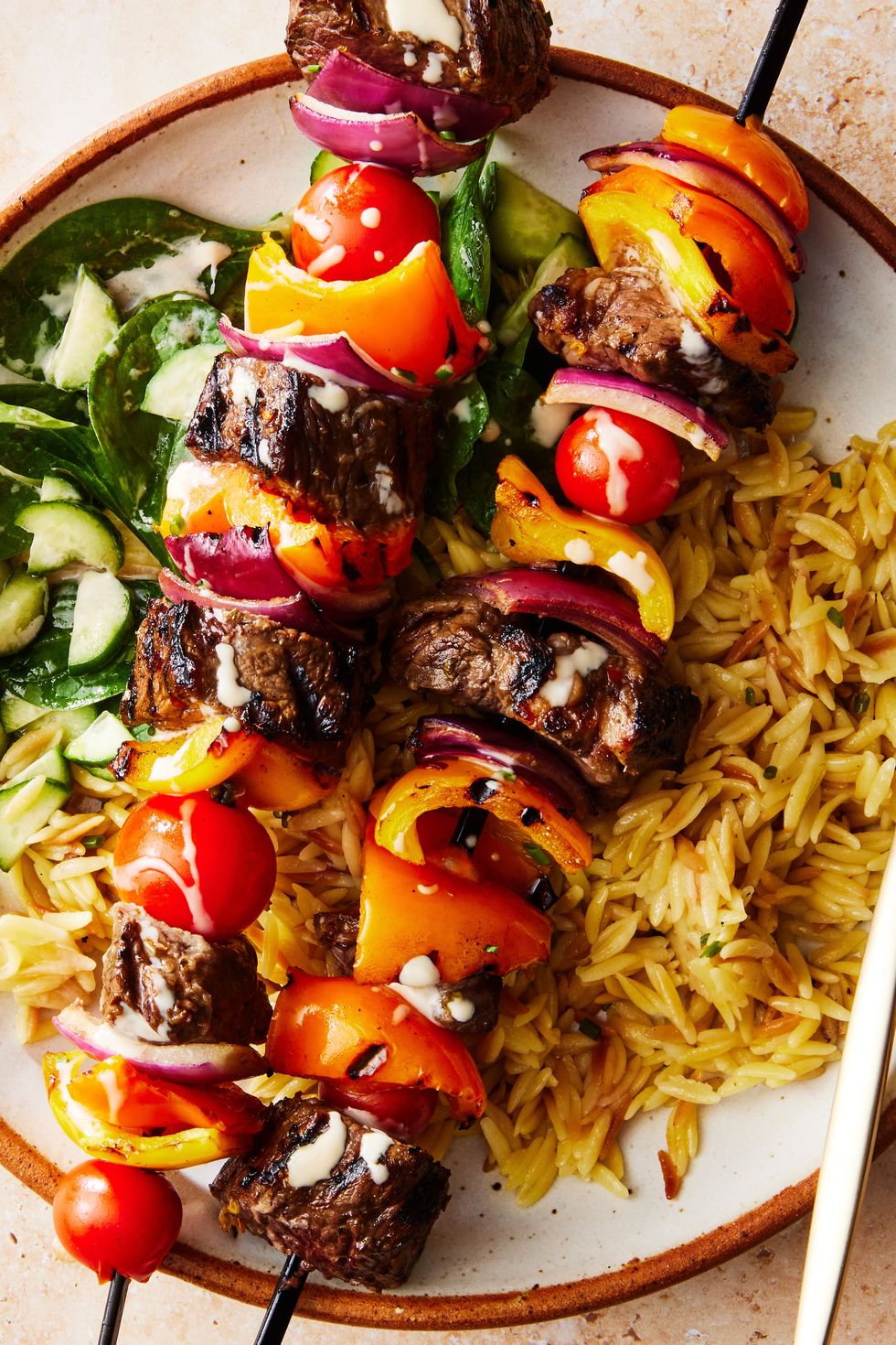 steak kebabs with peppers and tomatoes on top of orzo with a spinach and cucumber salad
