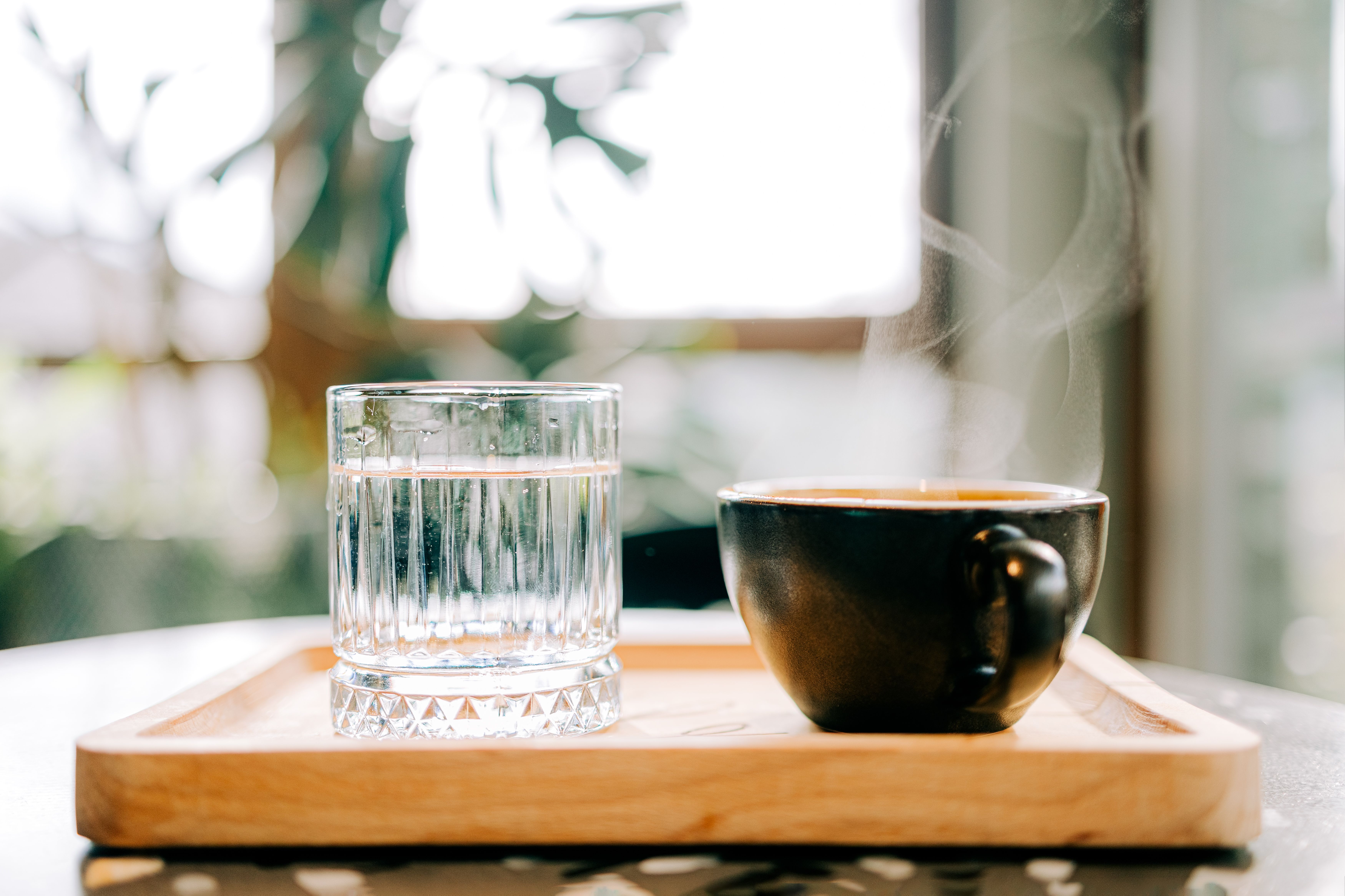 A glass of water and a steaming cup of black coffee on a wooden tray demonstrating how can black coffee count as water intake
