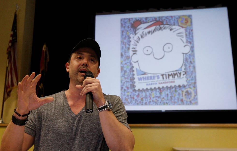 stephan pastis talking into a microphone, wearing a gray t shirt, and standing in front of a projected image of the cover of his children's book where's timmy