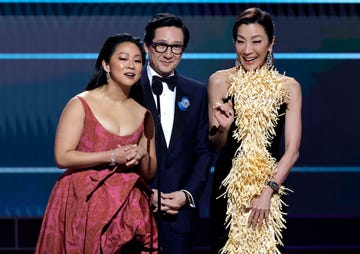 stephanie hsu, wearing a red dress, ke huy quan, wearing a black tuxedo, and michelle yeoh, wearing a yellow dress, smile and speak into a microphone on a stage during an awards ceremony
