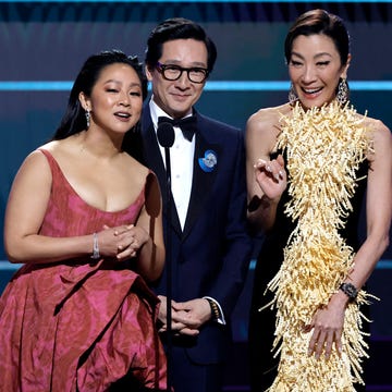 29th Annual Screen Actors Guild Awards - Show stephanie hsu, wearing a red dress, ke huy quan, wearing a black tuxedo, and michelle yeoh, wearing a yellow dress, smile and speak into a microphone on a stage during an awards ceremony