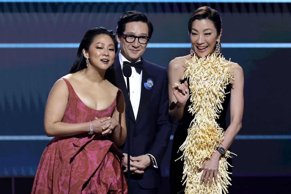 stephanie hsu, wearing a red dress, ke huy quan, wearing a black tuxedo, and michelle yeoh, wearing a yellow dress, smile and speak into a microphone on a stage during an awards ceremony