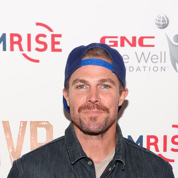 stephen amell stands looking at the camera with a neutral facial expression, he has brown hair and beard, he wears a cap backwards, grey tshirt, blue denim jacket and jeans