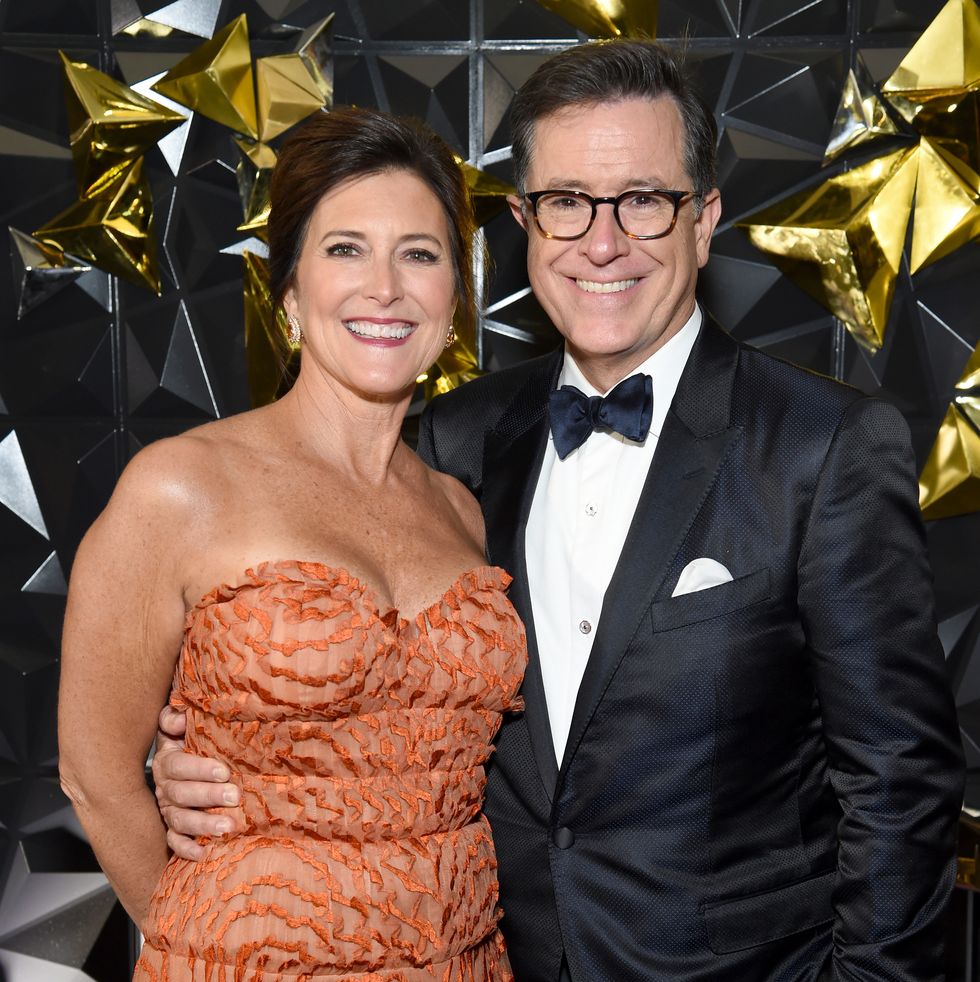 evelyn mcgee colbert and stephen colbert smile for a photo while standing together, she wears an orange strapless gown and he wears a black tuxedo with a bow tie and white pocket square