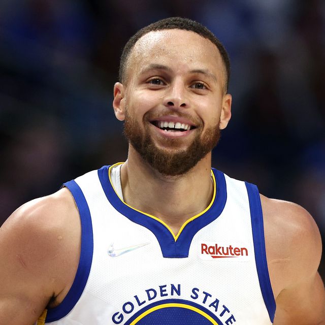 stephen curry smiles and looks past the camera, he wears a white golden state warriors jersey with blue and yellow accents