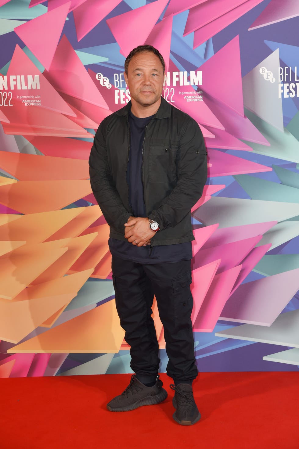 stephen graham, a man stands clasping hands and looking at the camera, he wears black trousers, tshirt, jacket and trainers