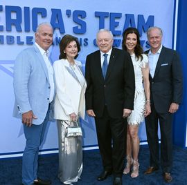 three men and two women stand together in a line on a blue carpet and pose for photos, behind them is a blue background that reads america's team