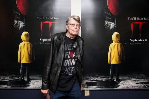 stephen king wearing a black t shirt, black leather jacket, and jeans, standing in front of posters for the movie it