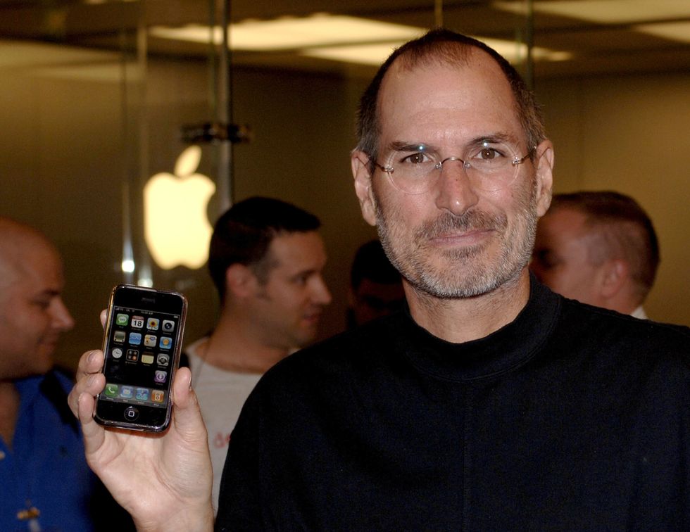 steve jobs smiling for a picture while holding an iphone with his right hand