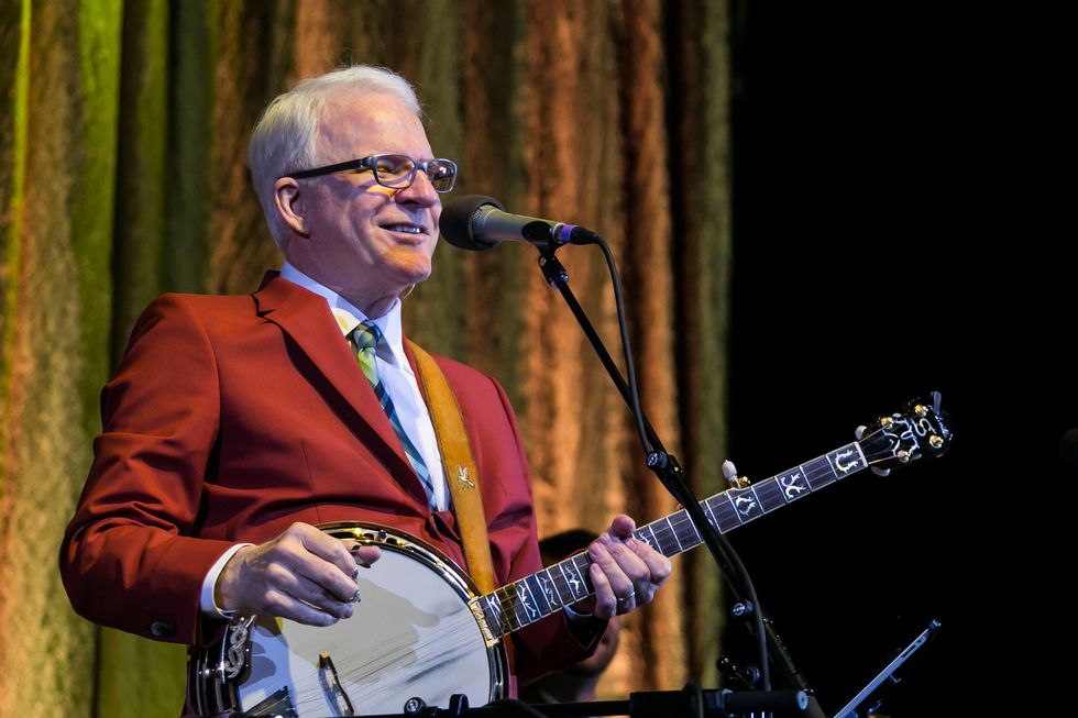 steve martin smiles while standing behind a microphone and holding a banjo