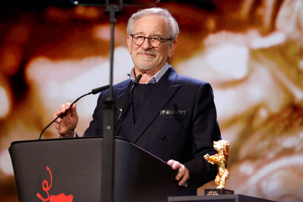 steven spielberg, wearing a black suit and glasses, speaks into a microphone at a podium, with the golden bear statue sitting on a table next to him