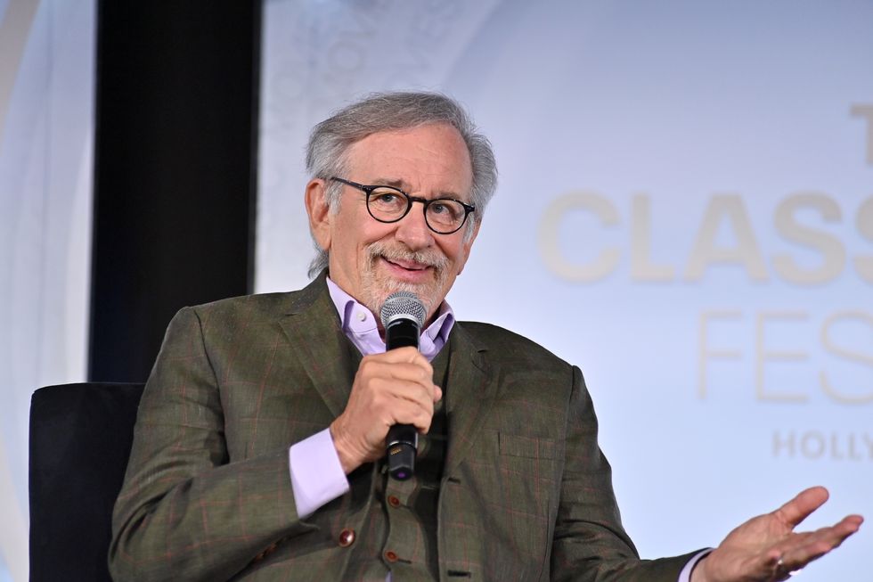 steven spielberg smiles as he speaks into a microphone he holds to his face, he is seated and has one hand extended to the right