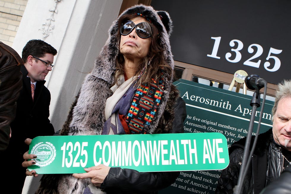 steven tyler, wearing a fur coat and sunglasses, holds a street sign for 1325 commonwealth avenue