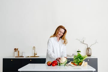 positive aged female in white shirt with long fair hair smiling and removing leaves from ripe cauliflower while preparing healthy lunch in modern kitchen at home
