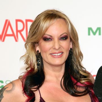 stormy daniels smiles at the camera while standing in front of a white backdrop with red and green text, she is wearing purple eye makeup, black dangling earrings, and a purple low cut gown decorated with a purple sequin pattern, her blonde hair is curled with sections of black and pink dyed hair, a tattoo on her right arm is partially visible