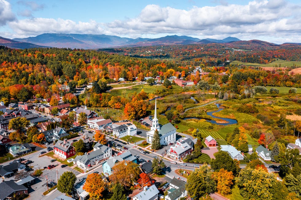 the scene captures the small town of stowe vermont with colorful autumn trees featuring a prominent white church steeple streets are lined with various buildings in shades of red, green, and blue a river meanders through green fields in the background with mountains rising in the distance under a partly cloudy sky