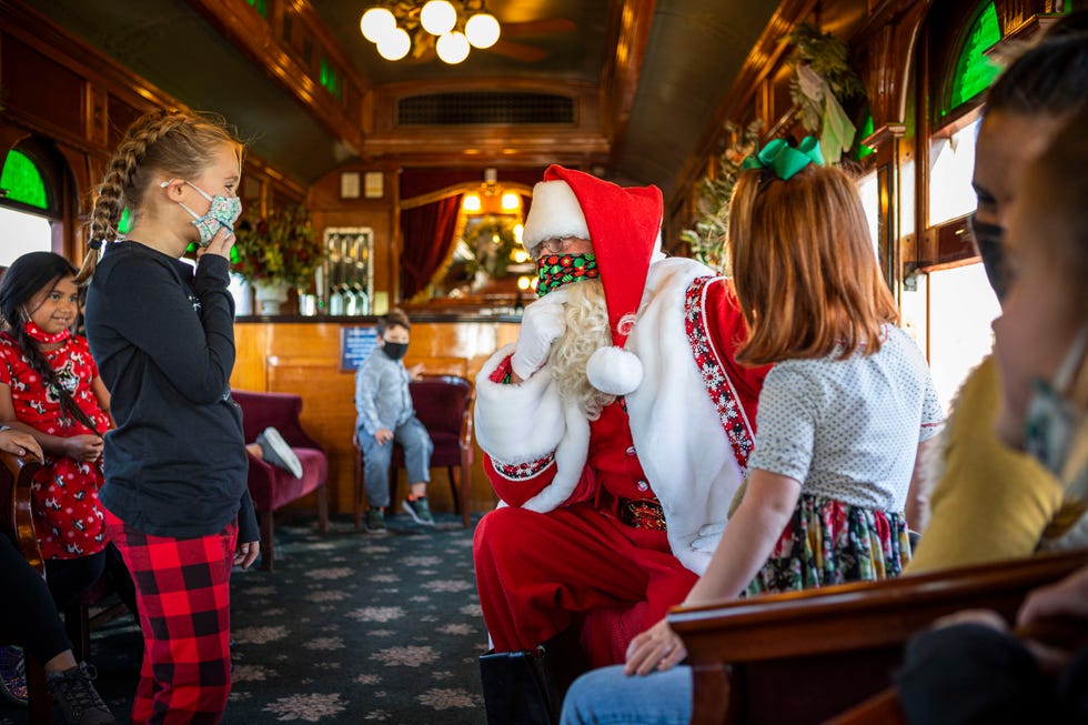 a masked santa is on an old fashioned train car kneeling in front of a little girl who is smiling behind her own mask there are other kids looking on