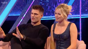 aljaz skorjanec and tasha ghouri on strictly it takes two a man wearing a black long sleeve shirt gestures with his hands while speaking, expressing a point next to him is a woman in a blue dress with a fitted bodice, listening attentively they are situated on a light colored couch, with a colorful, sparkling backdrop featuring blue and purple lighting