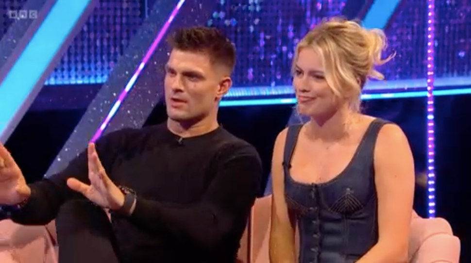 aljaz skorjanec and tasha ghouri strictly it takes two a man wearing a black long-sleeved shirt gestures with his hands as he speaks, expressing a point next to him is a woman in a blue dress with tight bodice, listening intently they are situated on a sofa light-colored, with a bright, colorful backdrop with blue and purple lighting