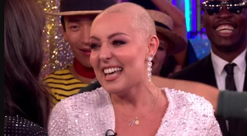 amy dowden smiles with people in the background, she has a bald head and wears diamonte drop earrings with a white sequin dress