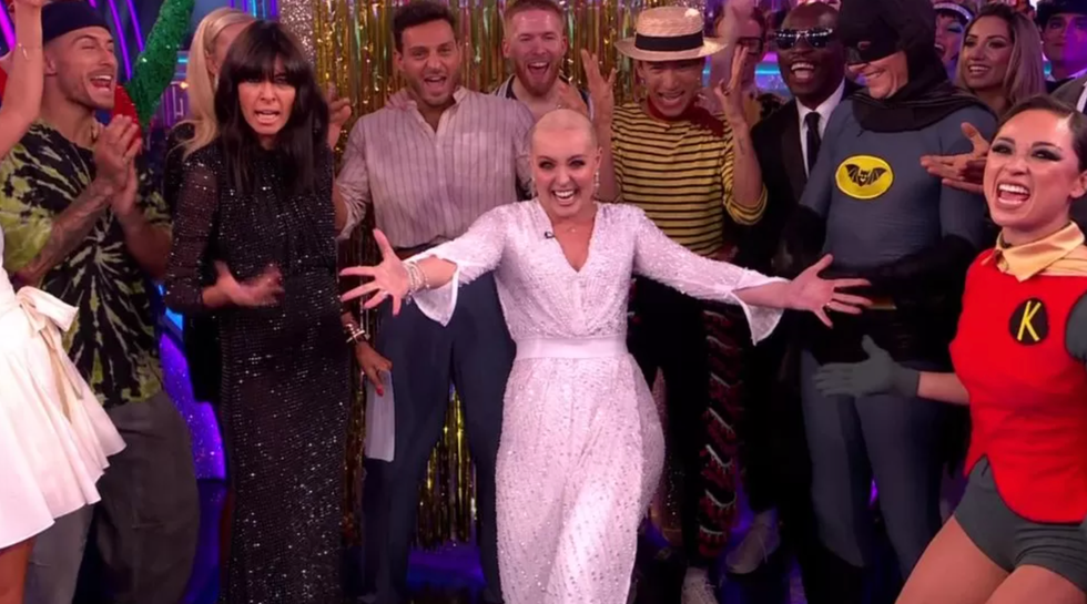amy dowden smiles with people in the background, she has a bald head and wears diamonte drop earrings with a white sequin dress