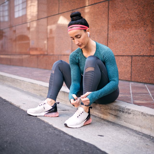 strong active woman sitting on the street sidewalk listening to music on wireless earbuds while tying her shoelace