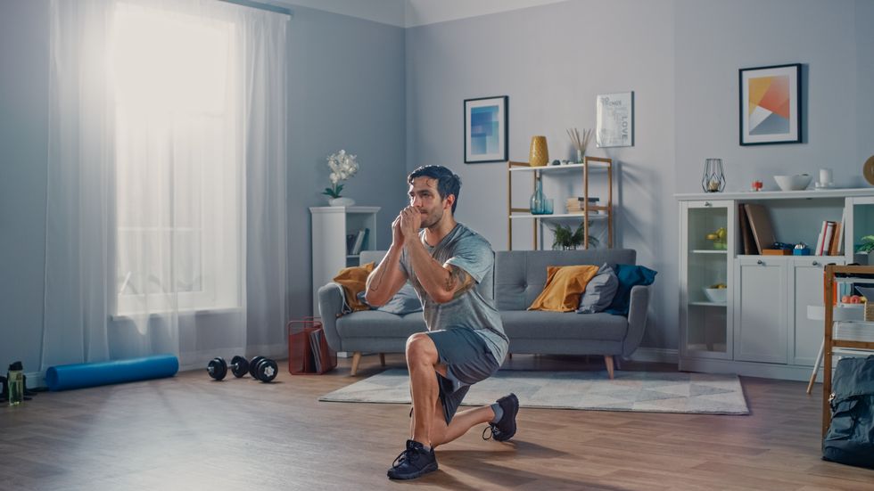 strong athletic fit man in t shirt and shorts is doing forward lunge exercises at home in his spacious and bright apartment with minimalistic interior