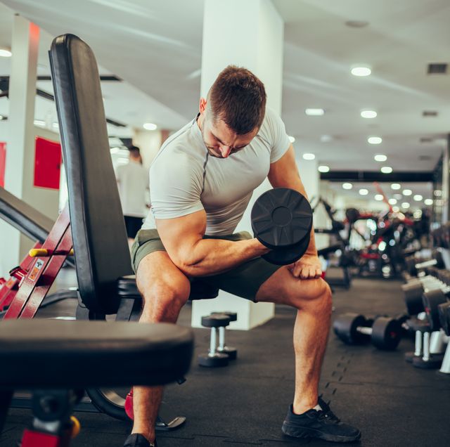 a strong sportsman is sitting in a gym next to the rack with dumbbells and exercising his biceps and triceps