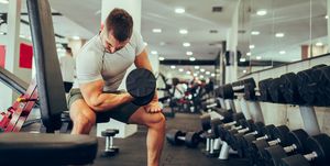 a strong sportsman is sitting in a gym next to the rack with dumbbells and exercising his biceps and triceps