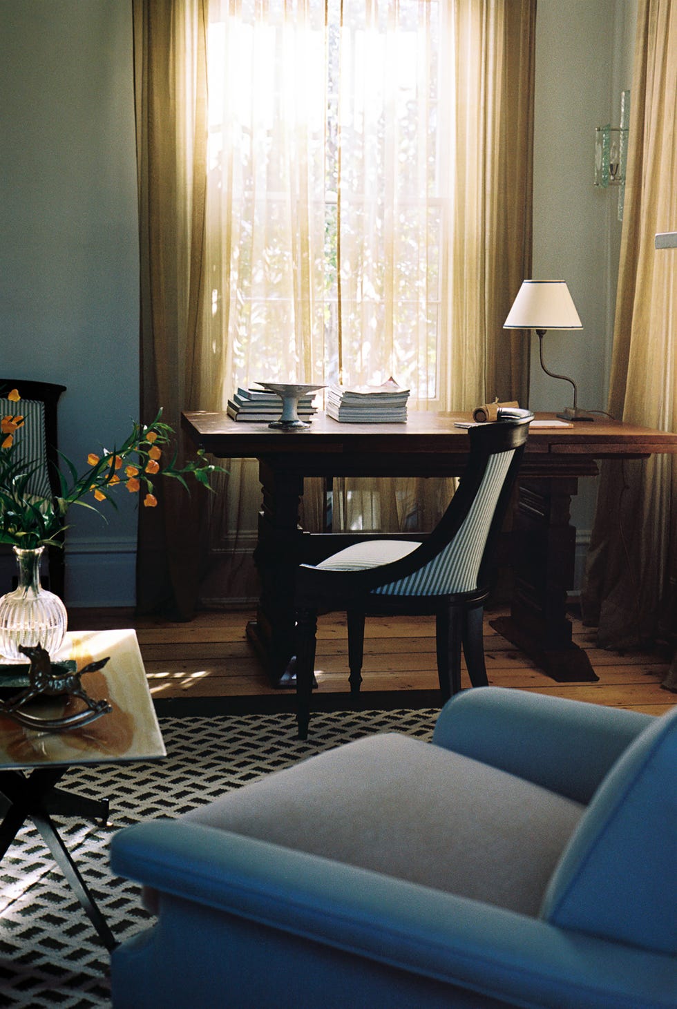 patterned rug underneath a blue armchair with a desk in the window and sun streaming through semi sheer curtains