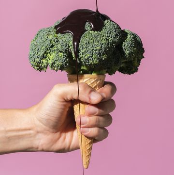 studio shot of man's hand holding ice cream cone with broccoli on top and pouring chocolate sauce, against pink background