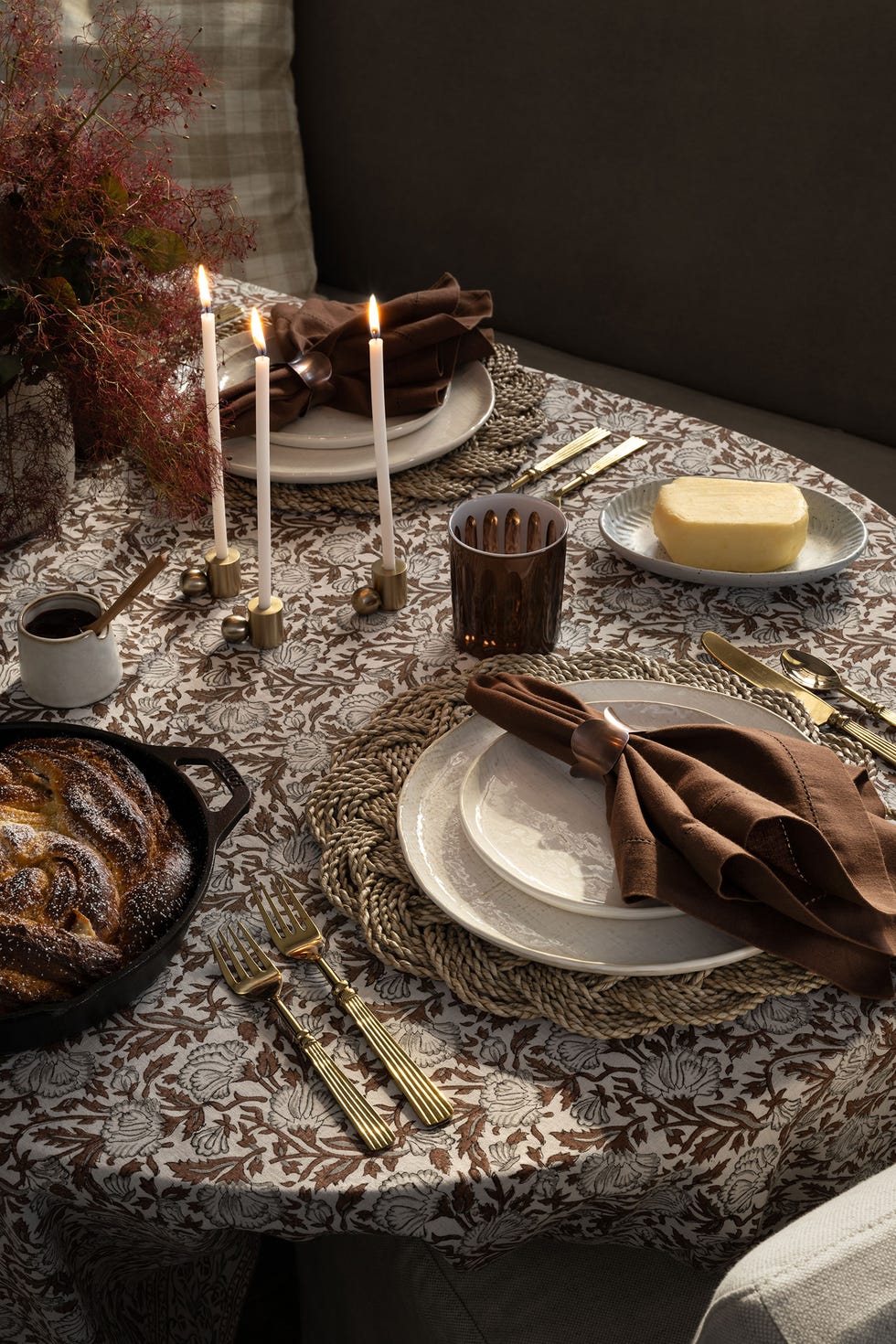 Thanksgiving Tablescape Ideas a beautifully arranged dining table featuring a patterned tablecloth three lit candles in gold holders sit at the center alongside decorative utensils several plates and bowls are placed, including a serving of butter, a small container of sauce, and a freshly baked pastry in a cast iron dish brown napkins are elegantly folded and held with napkin rings gold cutlery complements the sophisticated setting