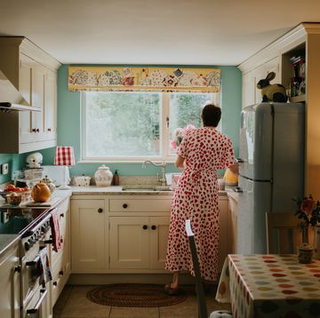 a stylish woman wearing a long printed dress stands at a sink in a small, rustic, country kitchen, and reaches for a kettle.
