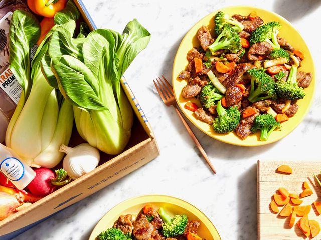 blue apron subscription services meal kit plated on marble table surrounded by vegetables in box and stirfry on yellow plates