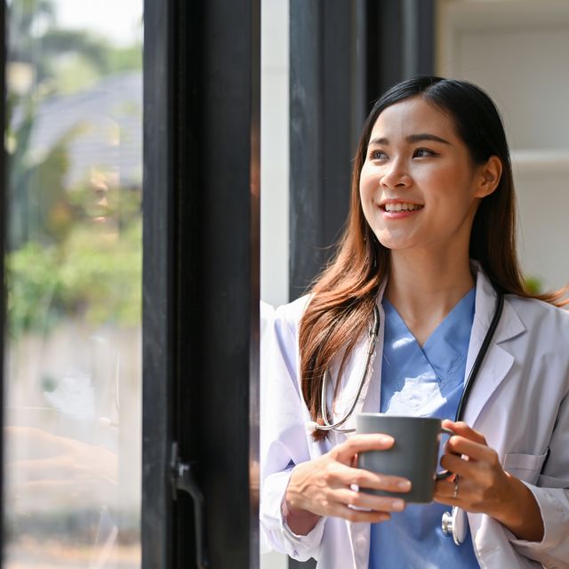 successful and inspired asian female doctor sipping coffee by the window in her hospital office