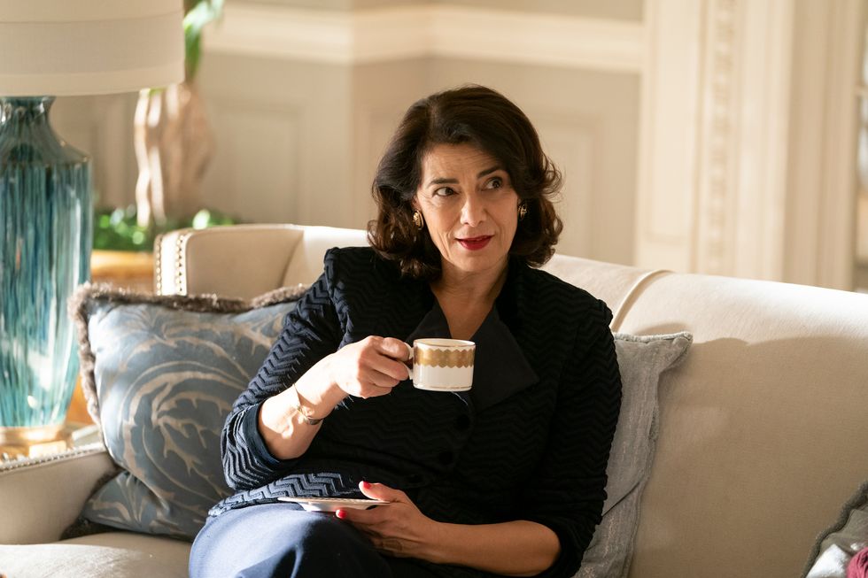 succession star hiam abbass, in character as marcia roy, sits on a sofa holding a cup and saucer in her hands