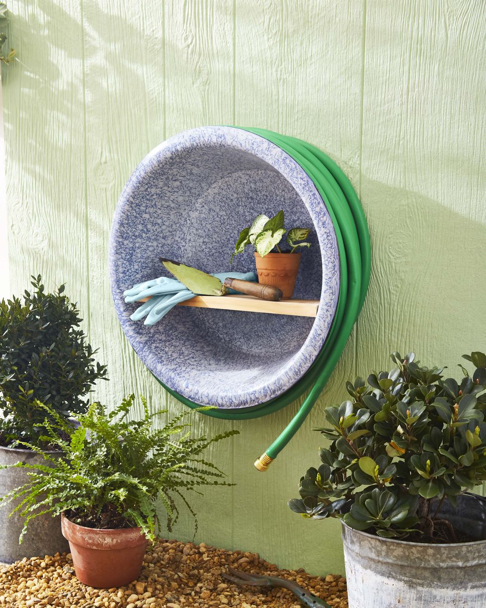 a summer garden caddy crafted from a large enamelware bowl mounted on a green exterior wall, with a hose wrapped around circumference and gloves and a spade stored on a wood shelf that spans the diameter
