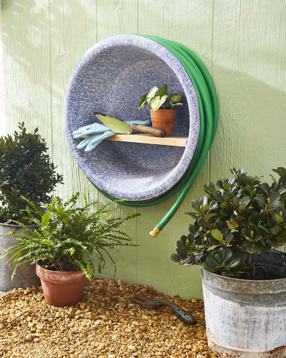 a summer garden caddy crafted from a large enamelware bowl mounted on a green exterior wall, with a hose wrapped around circumference and gloves and a spade stored on a wood shelf that spans the diameter