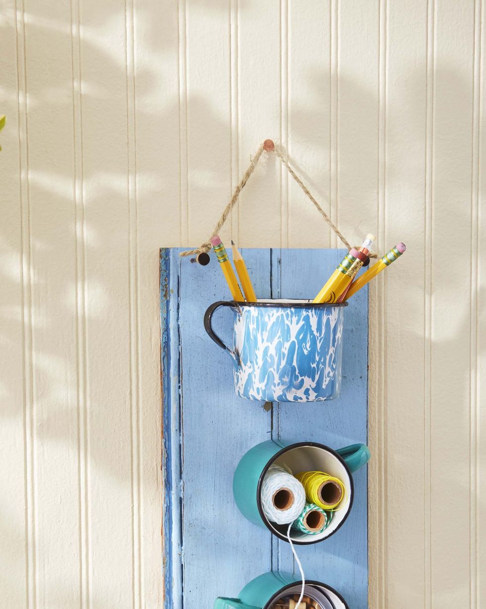 three enamelware mugs mounted on a piece of wood painted a summery blue serves as hanging storage piece for craft supplies, with pencils, spools of colorful thread, and push pins stashed in the mugs