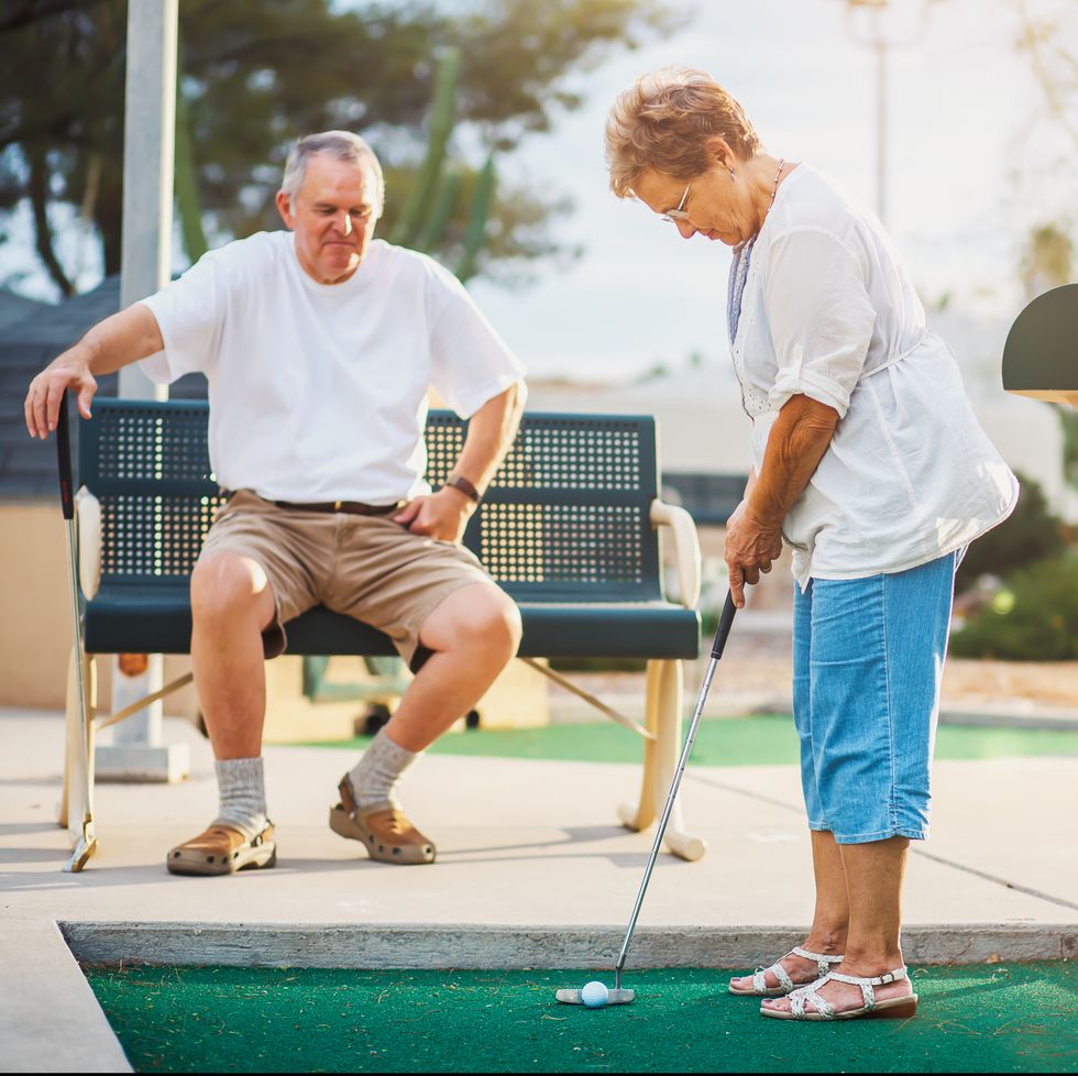 senior couple playing miniature golf, wife about to hit ball in hole while husband watches from bench on a summer date