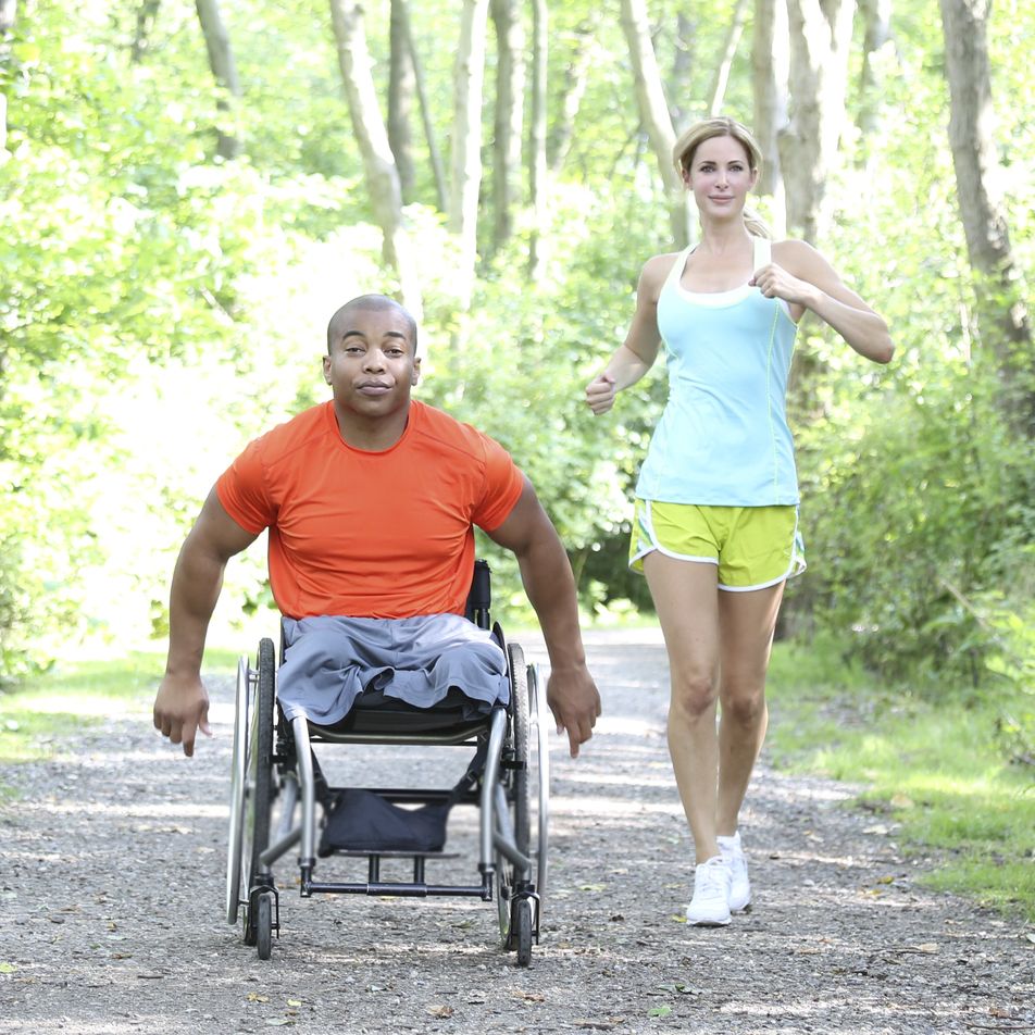 woman jogging behind man in wheelchair on outdoor trail on a summer date