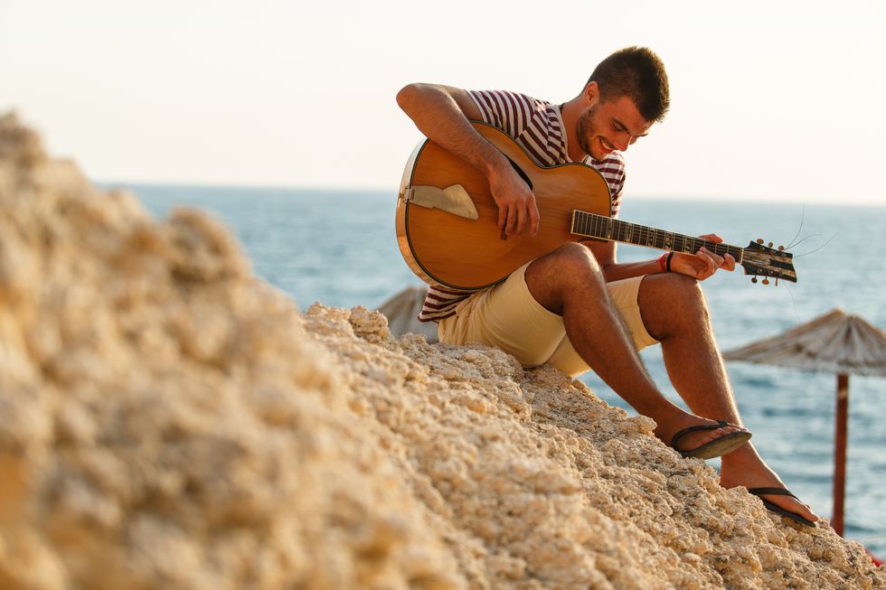 full length shot of joyful young man sitting on a rocky beach, enjoying summer sunshine and playing acoustic guitar