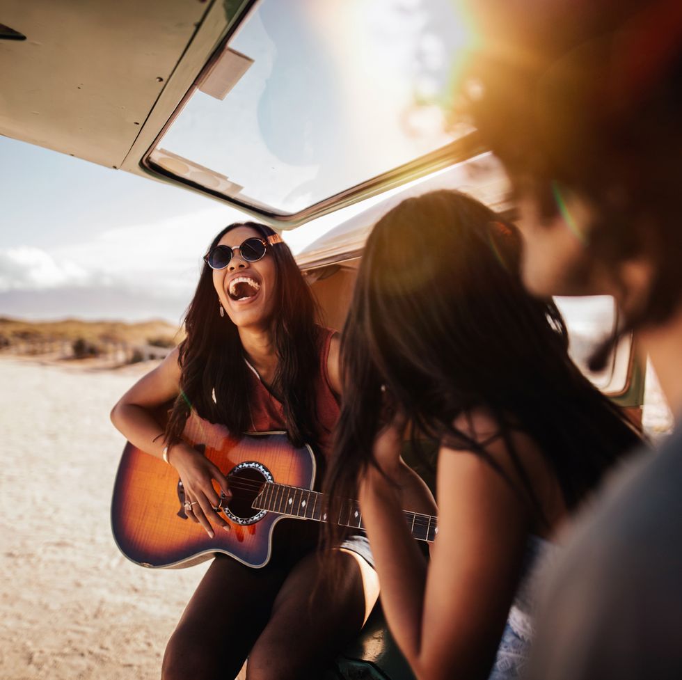 group of friends sit in back of open retro van parked at a sandy beach, playing guitar and smiling at each other on their road trip