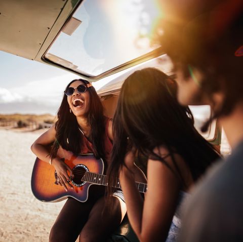 group of friends sit in back of open retro van parked at a sandy beach, playing guitar and smiling at each other on their road trip