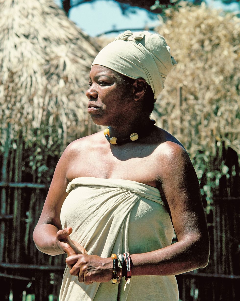 maya angelou stands in costume for roots, she wears a matching strapless wrap dress and turban and a beaded necklace, she stands and looks left in front of straw covered buildings
