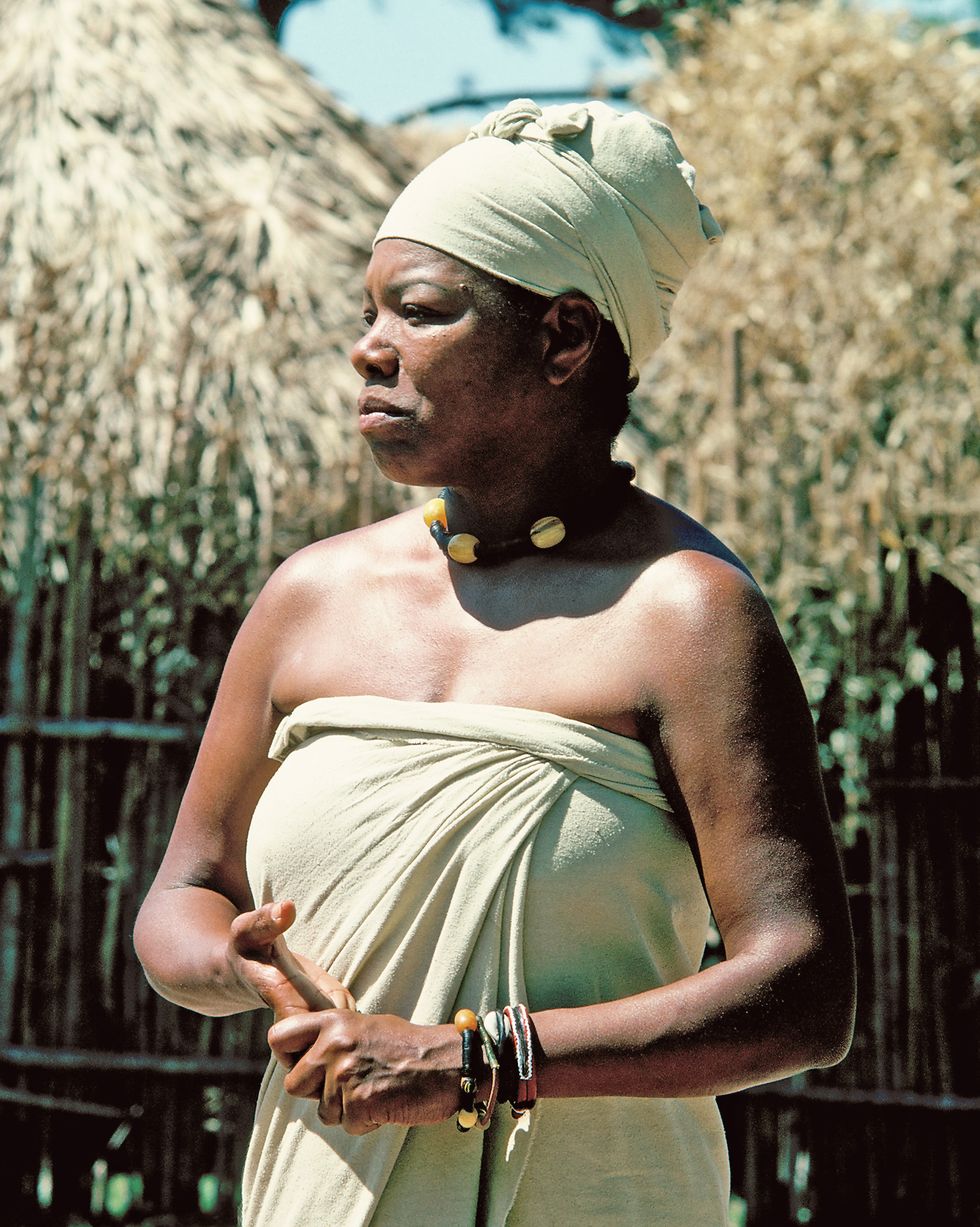 maya angelou stands in costume for roots, she wears a matching strapless wrap dress and turban and a beaded necklace, she stands and looks left in front of straw covered buildings