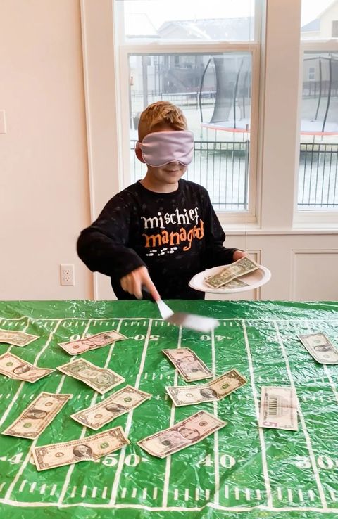 super bowl party ideas, boy with eyes covered holding a plate of money and a spatula with money on the table