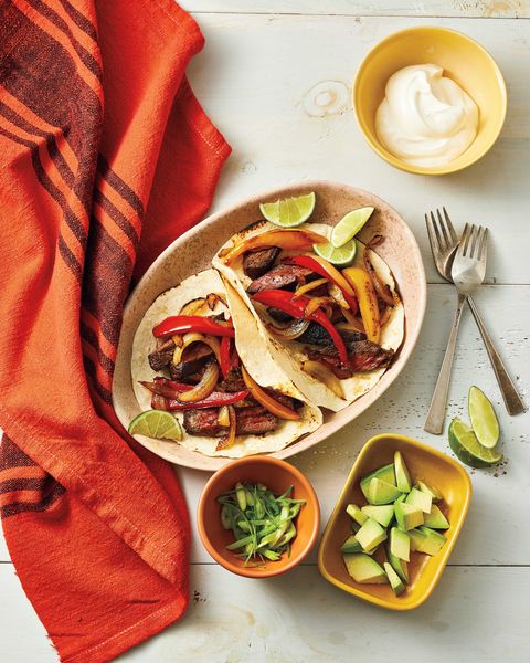 air fryer fajitas with lime wedges in a light tan colored oval bowl with small bowls of sliced green onion diced avocados and sour cream