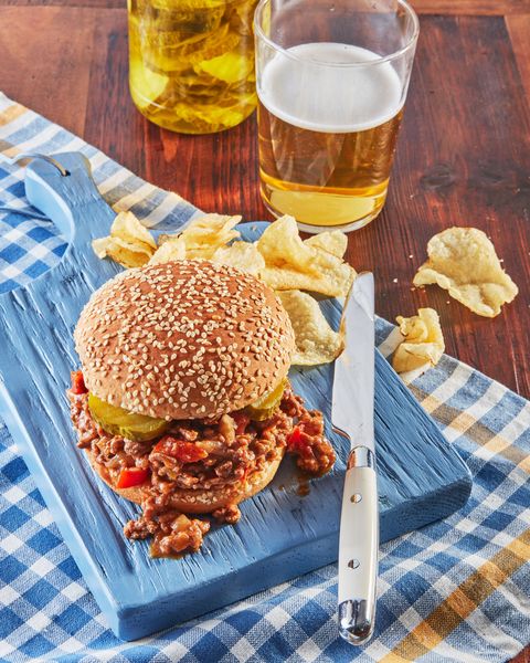 a sloppy joe with a sesame seed bun on a blue wooden serving board with a knife and chips
